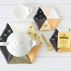 May include: A white ceramic teapot and a small white tea leaf holder sit atop a white wooden surface, surrounded by hexagonal fabric coasters. The coasters feature bee and honeycomb patterns in yellow, black, and cream colors. A gold spoon and a tea bag are also present.