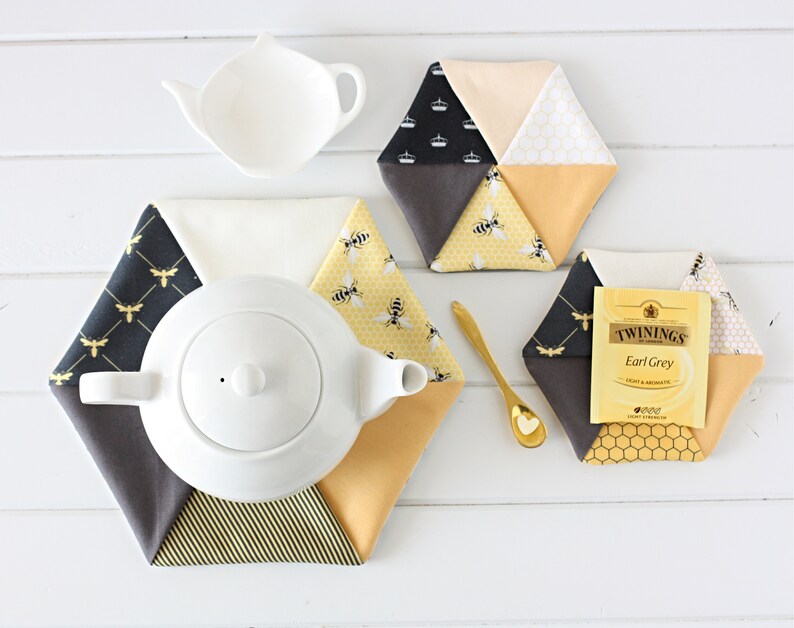 May include: A white ceramic teapot and a small white tea leaf holder sit atop a white wooden surface, surrounded by hexagonal fabric coasters. The coasters feature bee and honeycomb patterns in yellow, black, and cream colors. A gold spoon and a tea bag are also present.