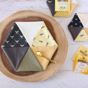 May include: Hexagon-shaped fabric coasters with bee and honeycomb patterns in yellow, black, and white. A small gold spoon with a heart-shaped detail rests on one coaster. Tea bags are also visible.