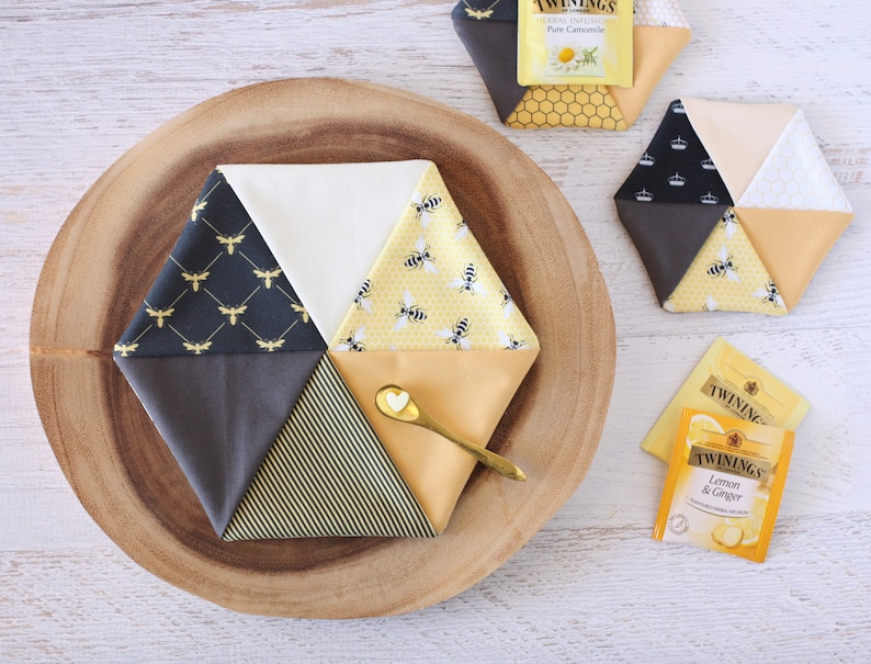 May include: Hexagon-shaped fabric coasters with bee and honeycomb patterns in yellow, black, and white. A small gold spoon with a heart-shaped detail rests on one coaster. Tea bags are also visible.