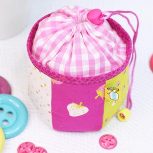 Pode incluir: Uma pequena bolsa com cord&atilde;o, com uma parte superior rosa e branca xadrez e um corpo multicolorido. O corpo apresenta pain&eacute;is rosa com um morango, branco com estrelas e amarelo com uma pequena etiqueta. Um cord&atilde;o e um fecho rosa prendem a bolsa.