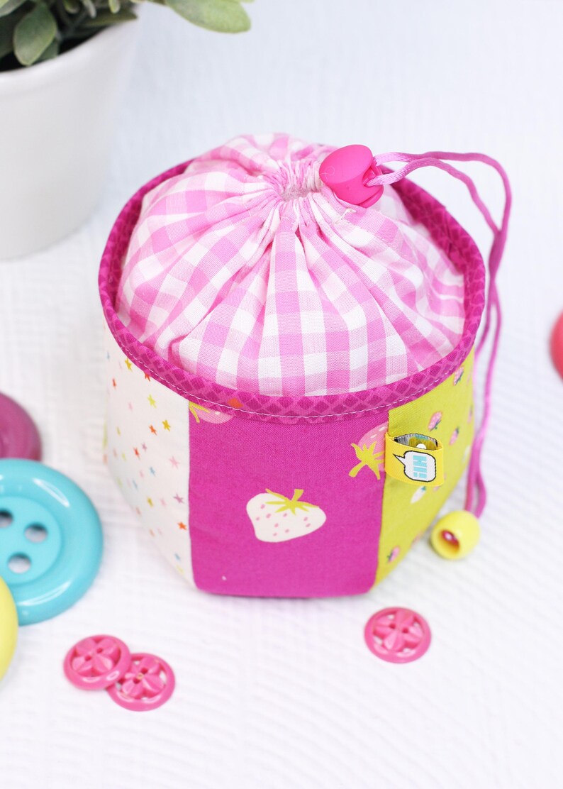 Pode incluir: Uma pequena bolsa com cord&atilde;o, com uma parte superior rosa e branca xadrez e um corpo multicolorido. O corpo apresenta pain&eacute;is rosa com um morango, branco com estrelas e amarelo com uma pequena etiqueta. Um cord&atilde;o e um fecho rosa prendem a bolsa.