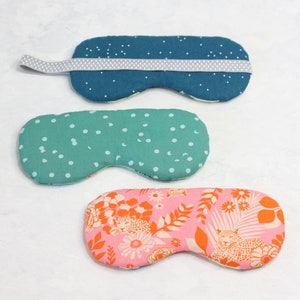 May include: Three sleep masks with different patterns. The first mask is blue with white polka dots and a gray elastic band. The second mask is green with white polka dots. The third mask is pink with a floral pattern and orange accents.