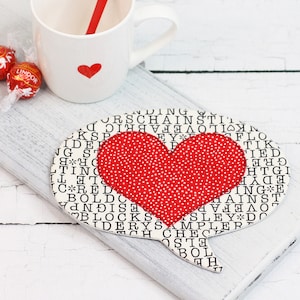 May include: A heart-shaped fabric coaster with a red polka-dot heart applique. The background is white with black text. A white mug with a red heart and a red straw is in the background.