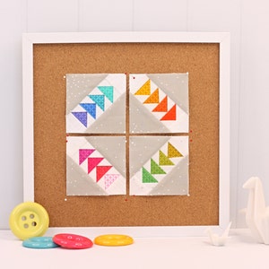 Puede incluir: Cuatro cuadrados de tela con un diseño de molinete en un esquema de colores arcoíris se muestran en un tablero de corcho con un marco blanco. Los cuadrados están dispuestos en una cuadrícula de 2x2.