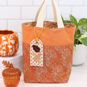 K&ouml;nnte beinhalten: Eine kleine orangefarbene und wei&szlig;e Tragetasche mit einem Stoffanh&auml;nger. Der Anh&auml;nger hat ein braunes Filzblatt und braune und wei&szlig;e Punkte. Die Tasche hat einen wei&szlig;en Griff und ist aus Stoff.