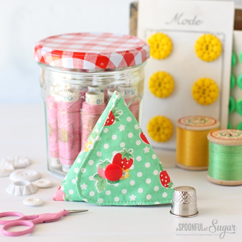 K&ouml;nnte beinhalten: Ein gr&uuml;nes und wei&szlig;es Stoffdreieck als N&auml;hnadel-Etui mit roten Erdbeeren und wei&szlig;en Punkten. Das Etui ist geschlossen und hat einen roten Knopf. Das Etui liegt auf einer wei&szlig;en Oberfl&auml;che neben einem silbernen Fingerhut und einer rosa Schere.