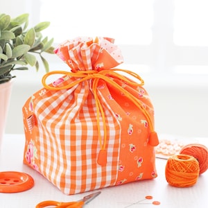 May include: An orange and white gingham drawstring bag with an orange drawstring. The bag is surrounded by sewing supplies, including orange thread, scissors, and a button. The bag has a floral pattern on the side.