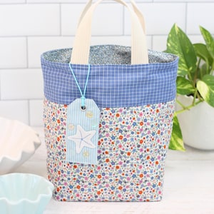 K&ouml;nnte beinhalten: Eine kleine blaue und wei&szlig;e Tragetasche mit Blumenmuster und einem blauen Anh&auml;nger mit einem wei&szlig;en Seestern. Die Tasche hat cremefarbene Henkel.