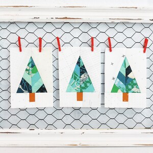 May include: Three handmade Christmas tree ornaments made from fabric scraps in shades of blue, green, and white. The ornaments are hung on a wire mesh background.