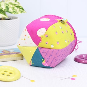 Pode incluir: Uma bola de tecido geom&eacute;trica colorida com estampas de morangos e estrelas. A bola &eacute; feita de v&aacute;rios pain&eacute;is de tecido em rosa, amarelo e azul-petr&oacute;leo. Um pequeno clipe amarelo est&aacute; preso. Outros itens de costura s&atilde;o vis&iacute;veis.