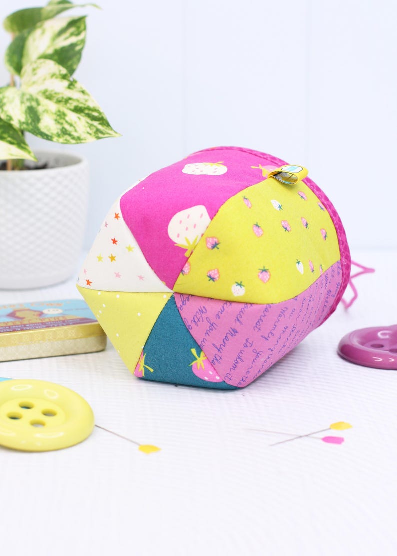 Pode incluir: Uma bola de tecido geom&eacute;trica colorida com estampas de morangos e estrelas. A bola &eacute; feita de v&aacute;rios pain&eacute;is de tecido em rosa, amarelo e azul-petr&oacute;leo. Um pequeno clipe amarelo est&aacute; preso. Outros itens de costura s&atilde;o vis&iacute;veis.