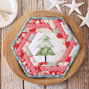 May include: A hexagonal quilted fabric mat featuring a central white panel with a green and white patterned Christmas tree design. The border consists of concentric strips of red, white, and light blue floral fabric with a blue patterned binding.