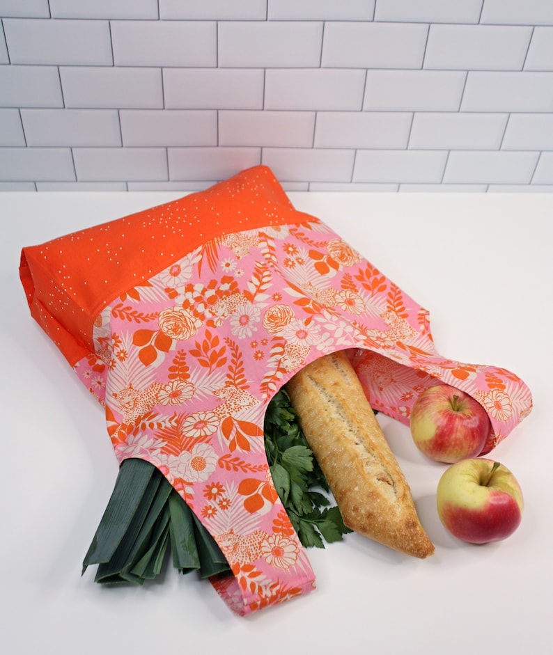 May include: A reusable shopping bag made of pink and orange fabric with a floral pattern. The bag is filled with groceries, including a baguette, leeks, and apples.