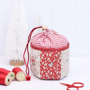 Pode incluir: Uma bolsa de tecido feita &agrave; m&atilde;o com uma parte superior xadrez vermelha e branca e um corpo de patchwork com padr&otilde;es florais e de p&aacute;ssaros. A bolsa tem um cord&atilde;o vermelho e um fecho de contas de madeira. Perto est&atilde;o bobinas de linha vermelha e tesouras.