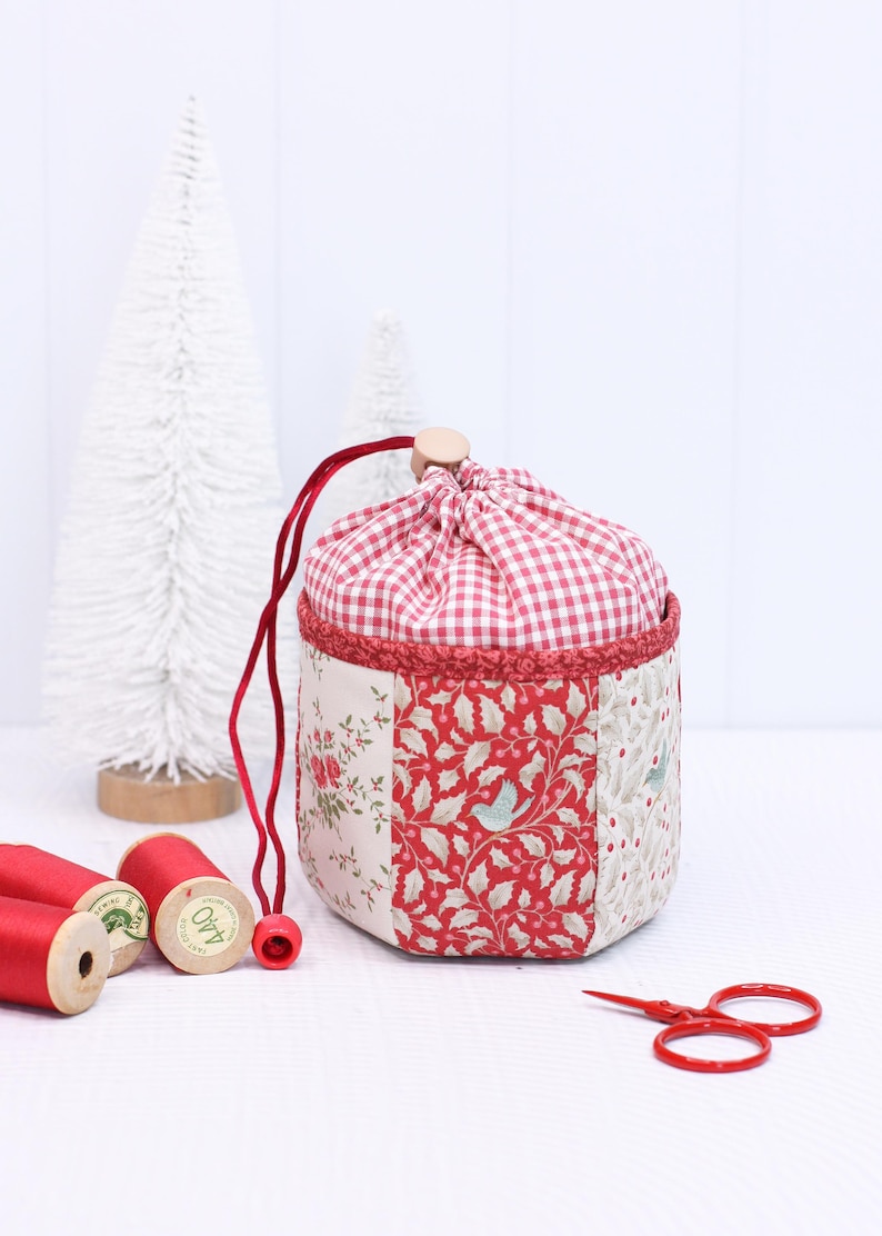Pode incluir: Uma bolsa de tecido feita &agrave; m&atilde;o com uma parte superior xadrez vermelha e branca e um corpo de patchwork com padr&otilde;es florais e de p&aacute;ssaros. A bolsa tem um cord&atilde;o vermelho e um fecho de contas de madeira. Perto est&atilde;o bobinas de linha vermelha e tesouras.