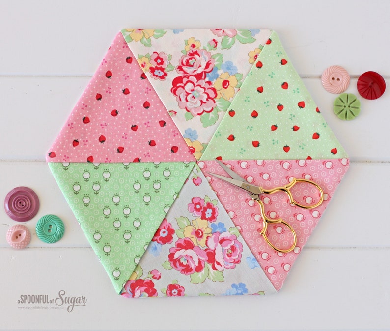 May include: A hexagonal patchwork sewing mat with various floral and strawberry patterns in pink, green, and white. Gold scissors rest on the mat, with several decorative buttons nearby. The mat is approximately 8 inches across.