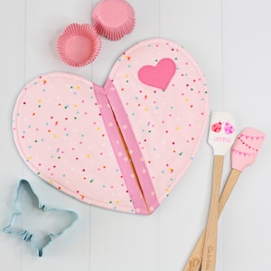 May include: Pink heart-shaped pot holder with a pink and white polka dot pattern and a pink heart applique. The pot holder is on a white surface with two pink cupcake liners and a blue butterfly cookie cutter.