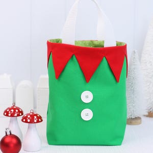 May include: A green fabric tote bag with a white handle and red trim, resembling an elf's outfit. The bag features two white buttons and a green interior with a polka dot pattern. Christmas-themed decorations are in the background.