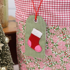 K&ouml;nnte beinhalten: Eine rote und wei&szlig;e Gingham-Stofftasche mit einer Stofftasche mit Blumendruck. Ein gr&uuml;nes Stoff-Geschenk-Etikett mit einem roten Filzsocken ist an der Tasche befestigt.