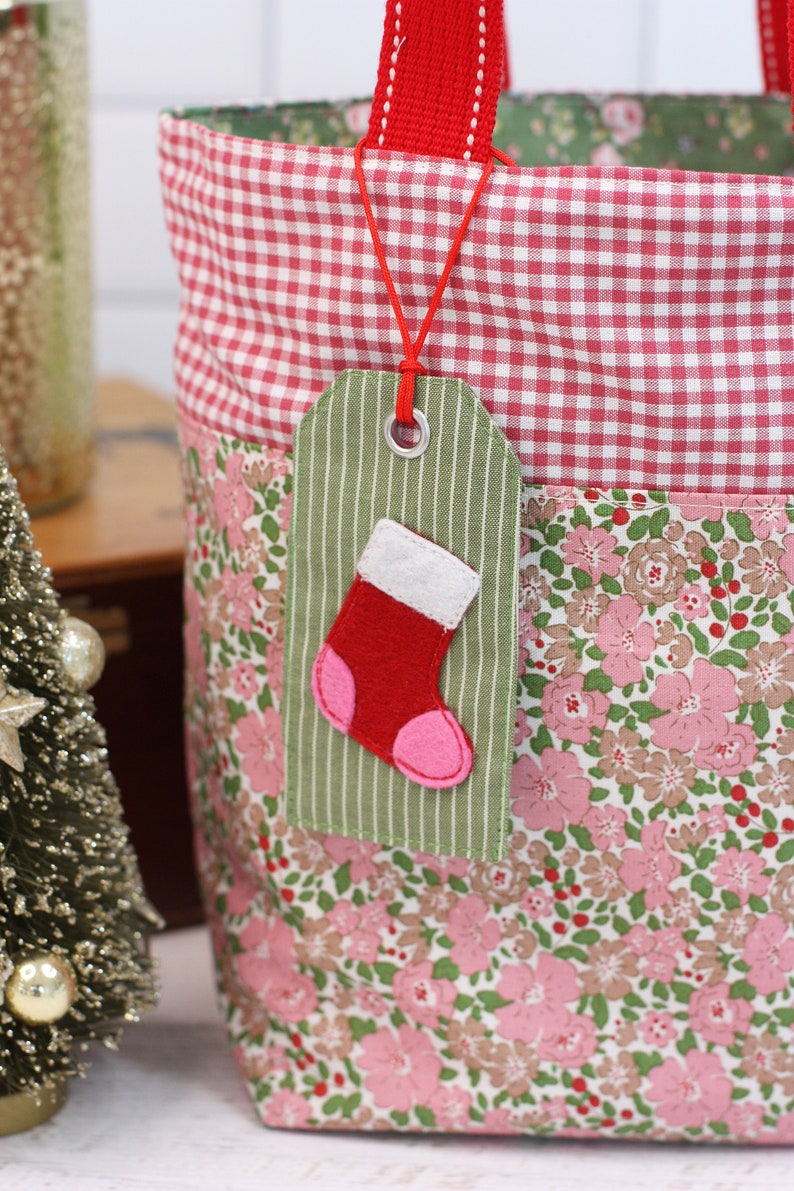 K&ouml;nnte beinhalten: Eine rote und wei&szlig;e Gingham-Stofftasche mit einer Stofftasche mit Blumendruck. Ein gr&uuml;nes Stoff-Geschenk-Etikett mit einem roten Filzsocken ist an der Tasche befestigt.