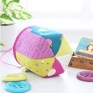 Pode incluir: Um estojo de costura de tecido geom&eacute;trico e colorido com um cord&atilde;o rosa. Apresenta pain&eacute;is estampados em verde-azulado, rosa, amarelo e branco, alguns com morangos e texto. Tr&ecirc;s bot&otilde;es amarelos, azuis e roxos em primeiro plano.