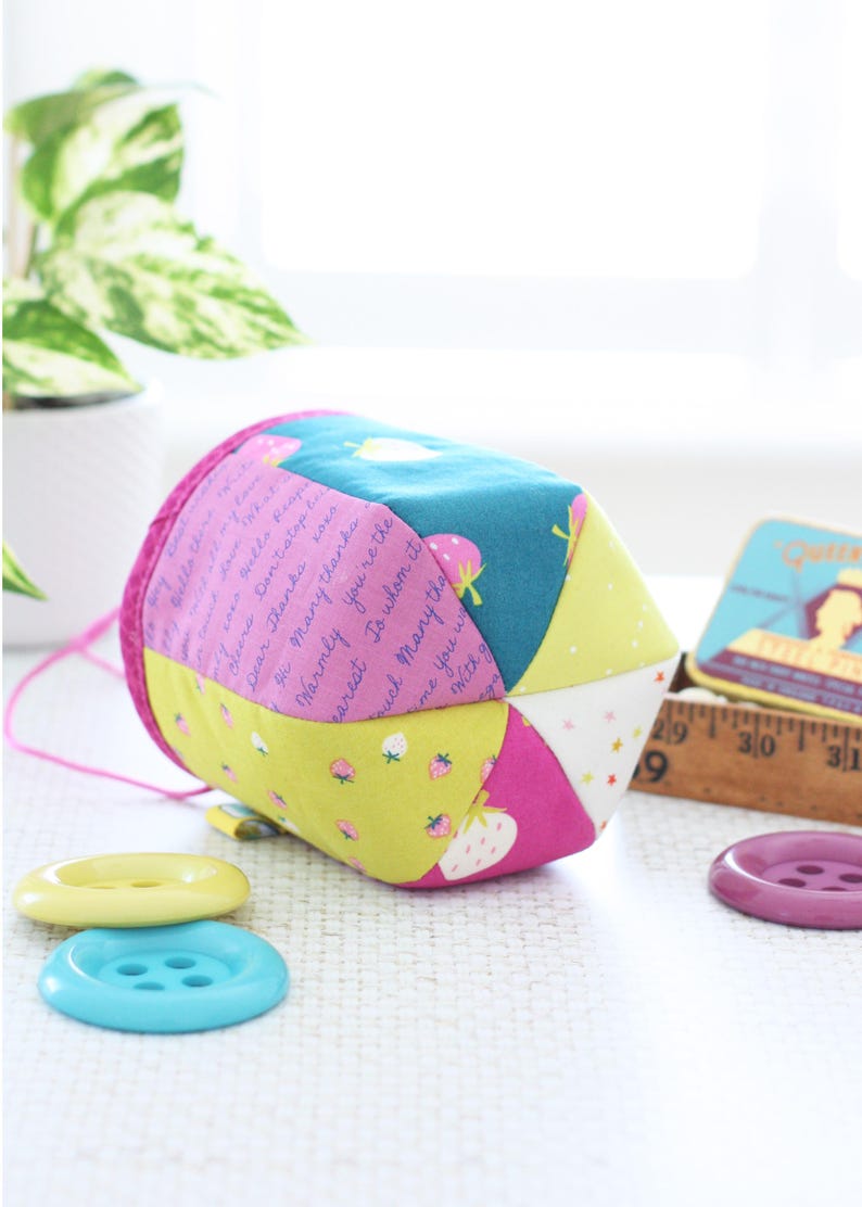 Pode incluir: Um estojo de costura de tecido geom&eacute;trico e colorido com um cord&atilde;o rosa. Apresenta pain&eacute;is estampados em verde-azulado, rosa, amarelo e branco, alguns com morangos e texto. Tr&ecirc;s bot&otilde;es amarelos, azuis e roxos em primeiro plano.