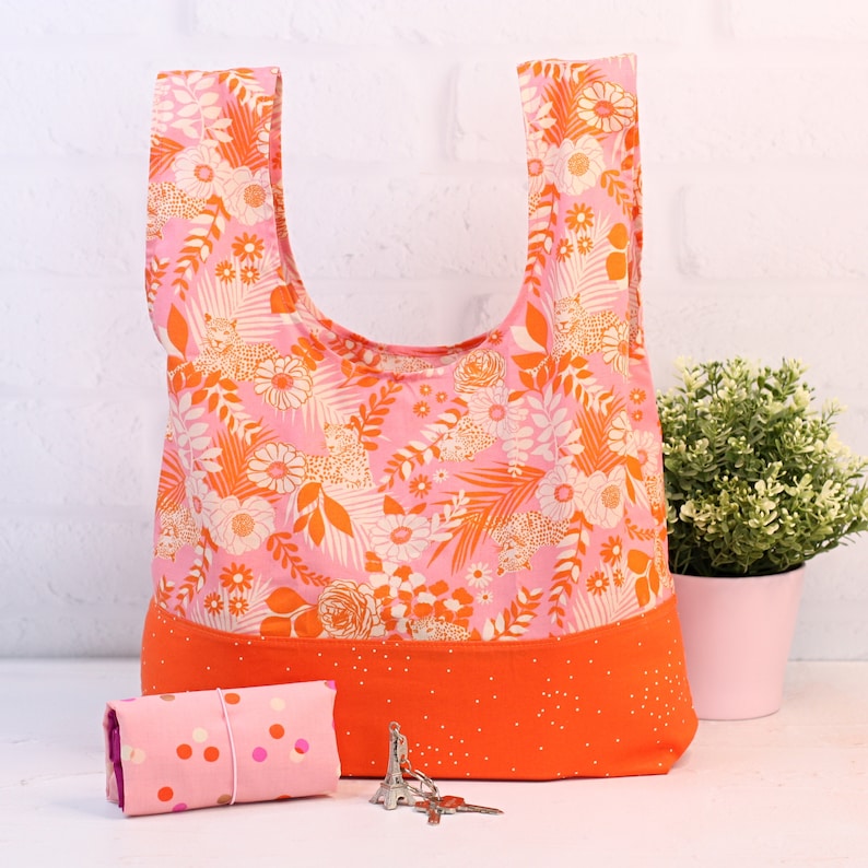 May include: A reusable shopping bag with a pink and orange floral print. The bag has a wide base and two long straps. The bag is folded and sitting on a white surface.