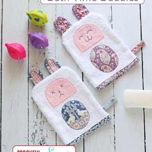 May include: Two white washcloths with embroidered bunny faces and floral fabric accents. The washcloths are on a white wooden surface with the text "Bath Time Buddies" and "a SPOONFUL of Sugar" printed above and below the washcloths.