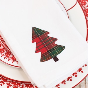 Peut inclure: Serviette en tissu blanc avec un motif d'arbre de Noël en tartan. La serviette est posée sur une assiette blanche avec un motif de flocons de neige rouges sur le bord. Décoration de table festive.