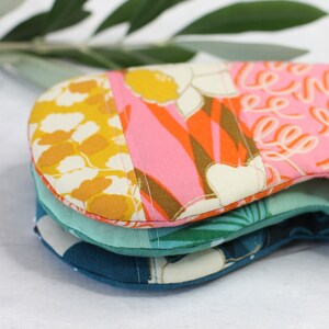 May include: Three eye masks made of fabric with different patterns. The top mask has a pink and orange floral pattern, the middle mask has a green and blue tropical pattern, and the bottom mask has a blue and white geometric pattern.