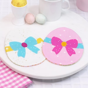 May include: Two fabric Easter egg-shaped coasters with bows. One coaster is white with a blue bow and gold glitter. The other coaster is pink with a pink bow and white snowflakes.