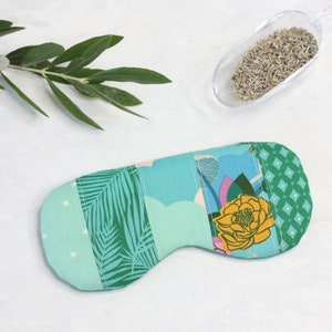 May include: A turquoise blue and green eye mask with a patchwork design featuring palm leaves, flowers, and geometric patterns. The eye mask is filled with lavender.