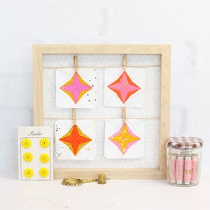 May include: Four colorful star-shaped fabric squares are displayed on a wooden frame with a white grid pattern. The squares are pink, orange, yellow, and red. The frame is hanging on a white wall. There is a small jar of pink thread spools and a button card with yellow buttons.