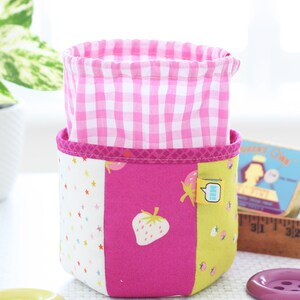 Pode incluir: Um cesto de armazenamento de tecido com interior rosa xadrez e exterior em patchwork. O exterior apresenta um painel branco com estrelas coloridas, um painel rosa com um morango e um painel amarelo com gr&aacute;ficos de morango. Uma pequena etiqueta est&aacute; anexada.