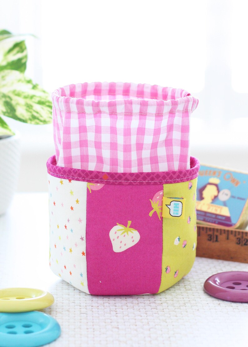 Pode incluir: Um cesto de armazenamento de tecido com interior rosa xadrez e exterior em patchwork. O exterior apresenta um painel branco com estrelas coloridas, um painel rosa com um morango e um painel amarelo com gr&aacute;ficos de morango. Uma pequena etiqueta est&aacute; anexada.