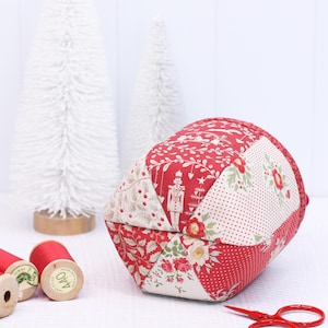 Pode incluir: Uma almofada de tecido patchwork em vermelho e creme com padr&otilde;es florais e de f&eacute;rias. A almofada est&aacute; perto de carret&eacute;is de linha vermelha, pequenas tesouras vermelhas e &aacute;rvores brancas.