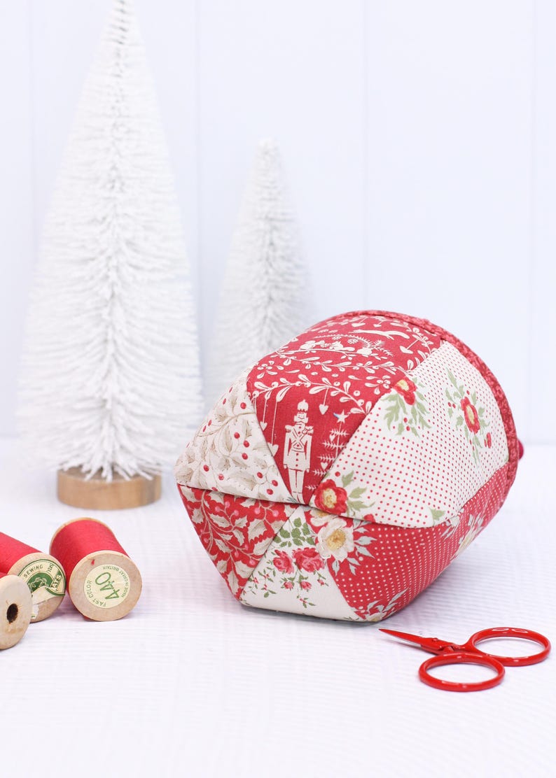 Pode incluir: Uma almofada de tecido patchwork em vermelho e creme com padr&otilde;es florais e de f&eacute;rias. A almofada est&aacute; perto de carret&eacute;is de linha vermelha, pequenas tesouras vermelhas e &aacute;rvores brancas.