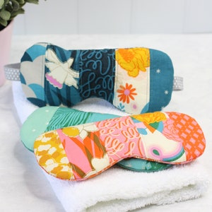 May include: Two patchwork fabric eye masks, one in shades of blue and white, and the other in pink, orange, and yellow, are lying on a white towel.