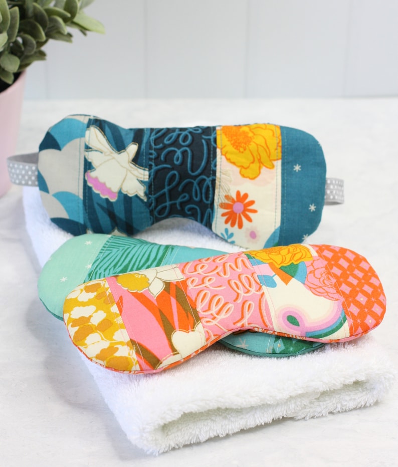 May include: Two patchwork fabric eye masks, one in shades of blue and white, and the other in pink, orange, and yellow, are lying on a white towel.