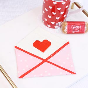 May include: A pink and white envelope with a red heart on the front. The envelope is on a white tray with a red mug and a package of Lotus Biscoff cookies.