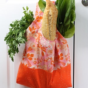 May include: A pink and orange reusable grocery bag with a floral pattern hangs on a white door. The bag is filled with a loaf of bread and leafy green vegetables.