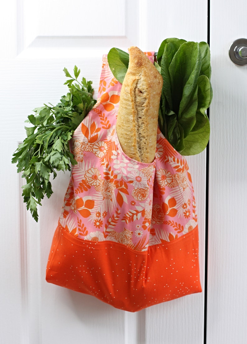 May include: A pink and orange reusable grocery bag with a floral pattern hangs on a white door. The bag is filled with a loaf of bread and leafy green vegetables.