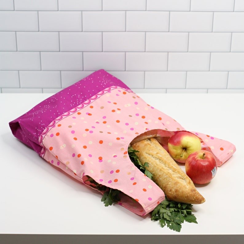 May include: A pink and purple reusable grocery bag with a polka dot pattern. The bag is filled with a loaf of bread, an apple, and a green leafy vegetable.