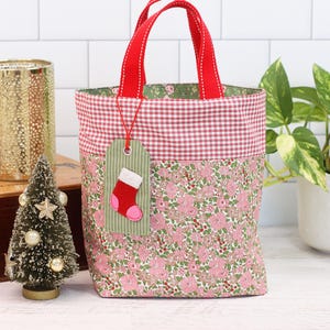 K&ouml;nnte beinhalten: Eine kleine rote und gr&uuml;ne Stofftasche mit roten Henkeln. Die Tasche hat ein Gingham-Muster auf der oberen H&auml;lfte und ein Blumenmuster auf der unteren H&auml;lfte. Ein gr&uuml;nes Etikett mit einem roten Filzsocken ist an der Tasche befestigt.