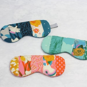 May include: Three colorful eye masks made of fabric with different patterns. The masks are all rectangular and have a strap to go around the head. The patterns include a blue and white floral design, a green and blue tropical design, and a pink and orange floral design.