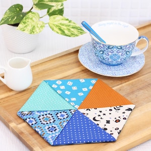 May include: A colorful, hexagonal fabric coaster with various patterns, including dice, polka dots, and geometric designs. A teacup and saucer with a blue floral pattern and a small creamer sit on a wooden tray, alongside a potted plant.