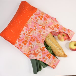 May include: A pink and orange reusable shopping bag with a floral pattern. The bag is filled with a loaf of bread, green onions, and apples.