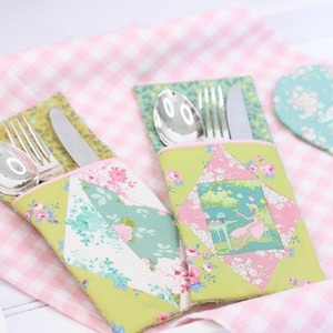 May include: Two quilted cutlery holders with floral and geometric patterns. Each holder contains a fork, knife, and spoon. The holders are green, pink, and white, with a pink and white gingham background.