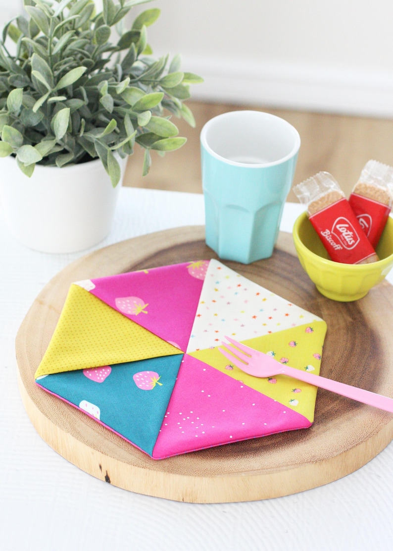 May include: A colorful, geometric fabric coaster with pink, yellow, and teal sections, adorned with strawberry and polka dot patterns. A pink fork rests on the coaster, alongside a teal cup and a small bowl with cookies.